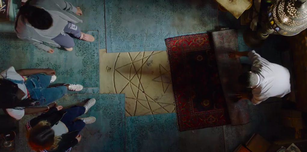 An overhead shot of four people standing around a Seal of Solomon carved into a floor covered in small, ornate blue rugs. A larger red Oriental-style rug is half-folded and being pulled away from the Seal.