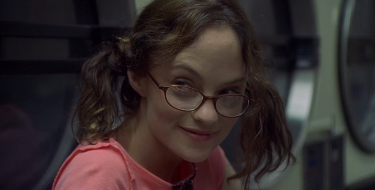 May looking off to the side, smiling slightly. She has kinda messy pigtails, dark-rimmed glasses, and a pink t-shirt.