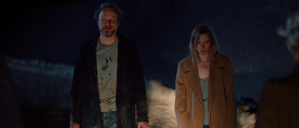 Bjørn and Louise standing in front of a rocky background, lit by car headlights. Bjørn has a bloody nose and bloody spatters on his shirt and has a sad, pained smile on his face. Louise has bruises on her nose and chest and she is looking downward with a blank expression.