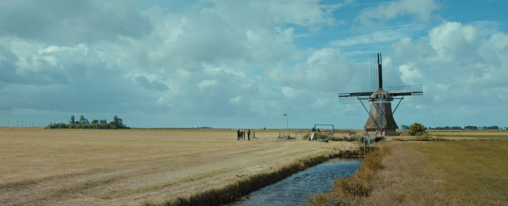 a wide shot of the Dutch landscape. The sky is very bright despite large clouds. There is a windmill with a canal running toward it. The two families are visible near the windmill. There is a lamp post, a soccer (football) goal, and a small slide. Abel is sitting at the bottom of the slide, and Agnes at the top.