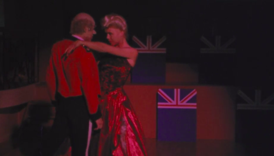 Tilbury and Gudrun dancing in a dark ballroom with small British half-flags hanging behind them. Tilbury is in a dress uniform and Gudrun is in a sparkly dark red gown.