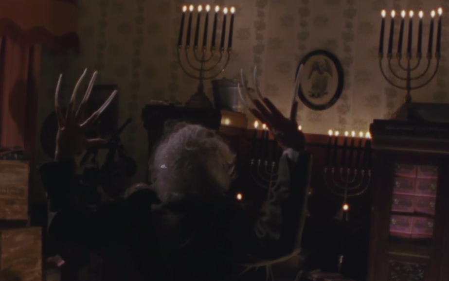 The tilberi seen from behind. His arms are stretched above his head and his fingers end in long claws. He's standing in front of some large shelves with four fully-lit menorahs on them.