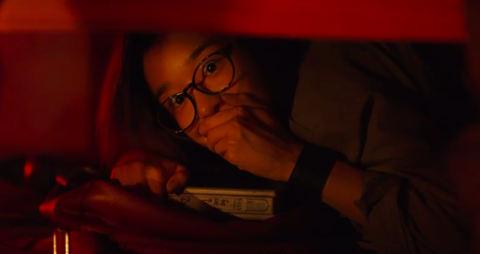 Mi-Jung hiding underneath a bed. She is a young Korean woman with thick-rimmed glasses, a dark green jacket, and a large, dark wristband. Her eyes are wide with fear and she's clasping her hand over her mouth. She's holding a VHS case in her other hand and the whole screen is tinted red.
