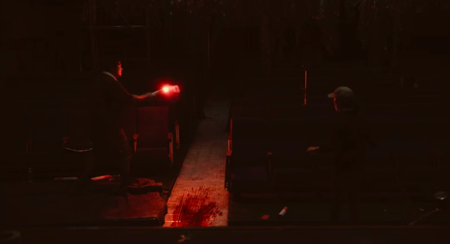 Two members of Jae-hyun's crew. They're in the center aisle of a very dark theater and the person on the left is holding a lit red flare. There's a large bloody smear on the ground.